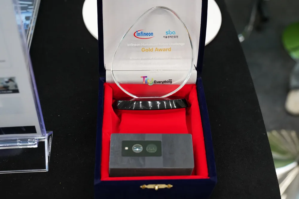 A ModigenceVision camera module in a presentation box beside the Infineon Humanoid Robot Challenge Gold Award trophy on the booth table
